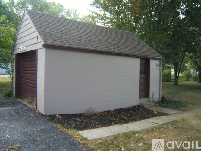 A small garage with a brown door is surrounded by a gravel driveway and a grassy area.