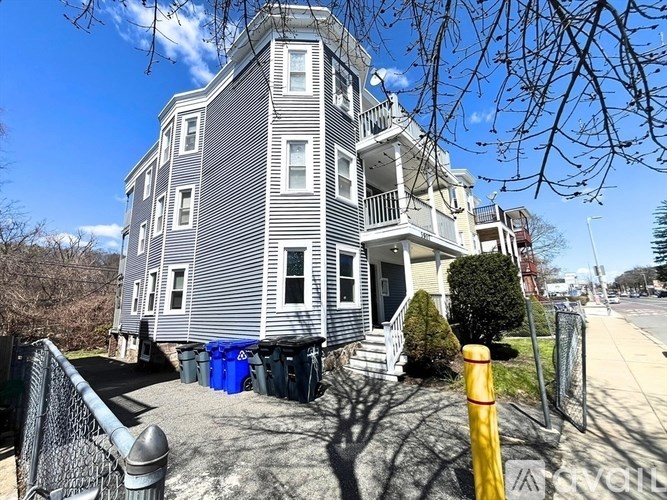 A grey house with a blue trash bin in front.