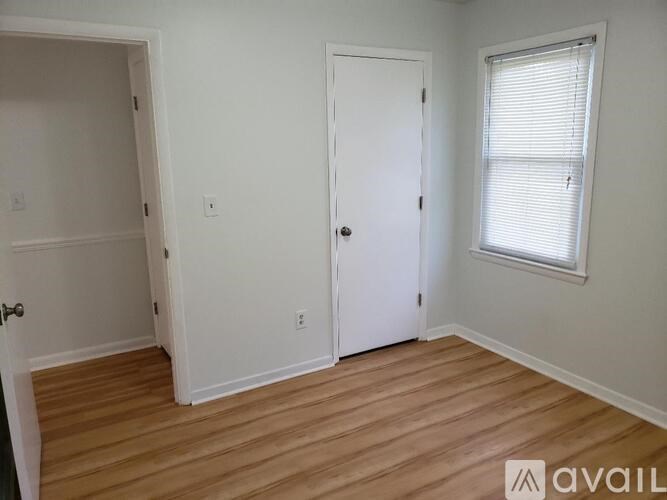 A room with a white door, a window with blinds, and a wooden floor.