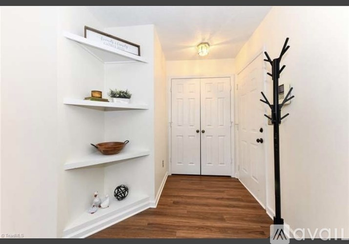 A hallway with a white wall, a wooden floor, a white door, a white shelf with a plant and candle, a black pole, and a black and white decoration.