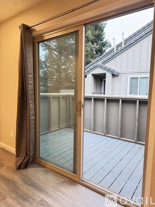 A sliding glass door with a curtain on the side opens to a deck with a house in the background.
