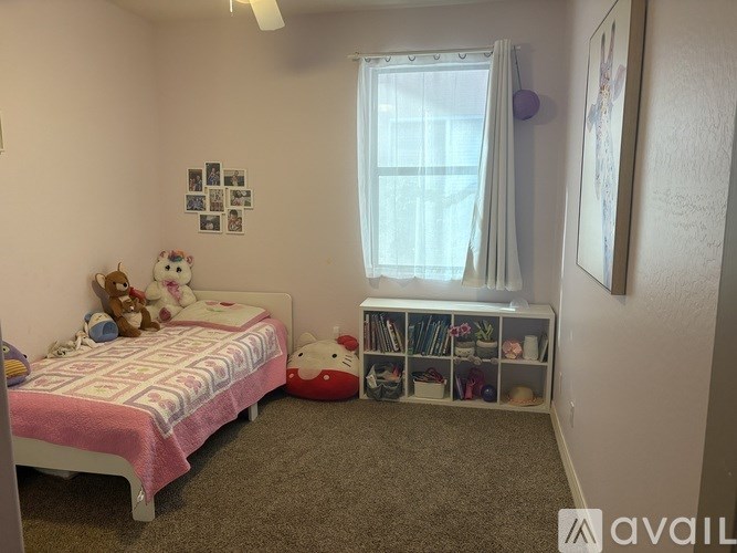 A small bedroom with a bed, a window with curtains, a shelf with books and toys, and pictures on the wall.
