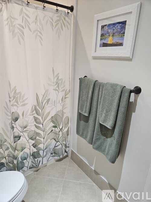 A bathroom with a toilet, a shower curtain with a leaf pattern, a towel rack with two towels, and a picture on the wall.