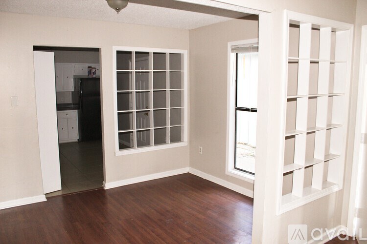 A room with a kitchen in the background and a window with blinds.