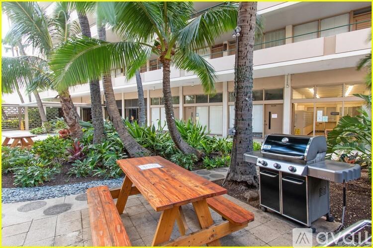 A wooden picnic table is in front of a grill and a building with a balcony.
