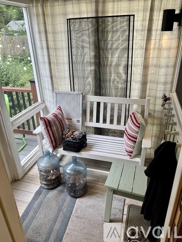 A white bench with red and white pillows sits in front of a window with curtains.