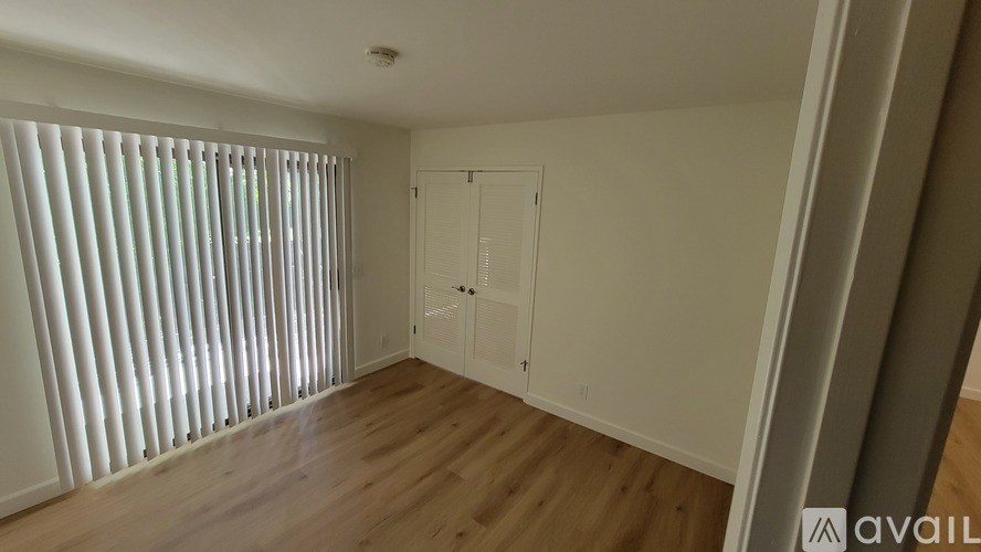 A room with wooden floors and white walls, featuring a window with blinds and a white door.