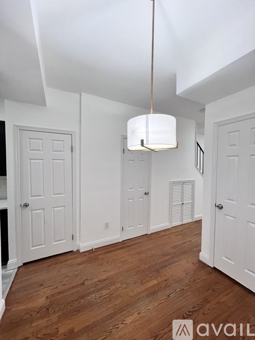 A room with white walls and wooden floors, with a hanging light fixture and a door on the left.