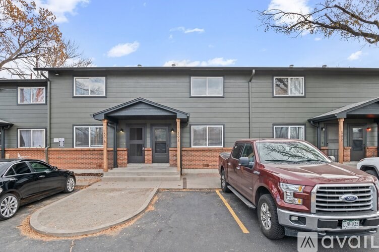 A red Ford truck is parked in front of a grey building with a black car to its left.
