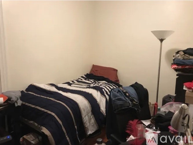 A bedroom with a bed covered in clothes and a lamp on the floor.