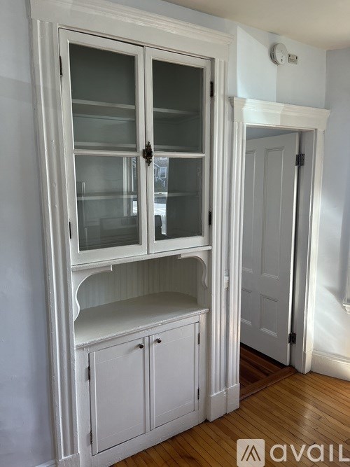 A white cabinet with glass doors and a shelf is in a room with wooden floors.