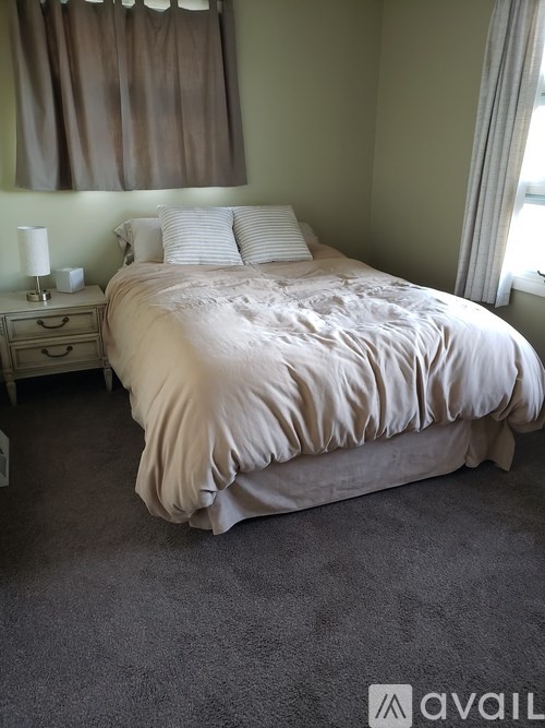 A bed with a white comforter and two white pillows sits in a room with a window and a nightstand with a lamp.