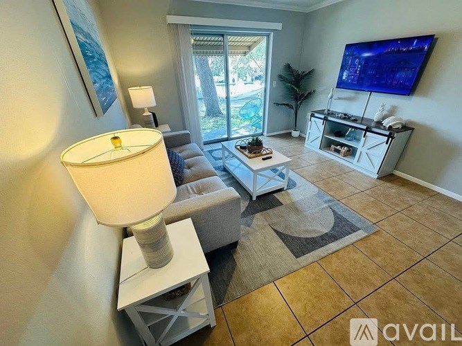 A living room with a grey couch, a white side table with a lamp, a coffee table, and a flat screen TV mounted on the wall.