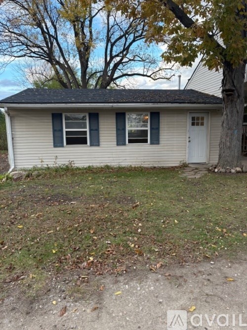 A small house with a white door and two windows is surrounded by a grassy area with fallen leaves.