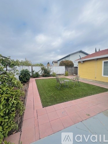 A backyard with a yellow house, a green lawn, and a red brick walkway.