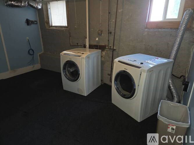 Two white front load washing machines in a room with a window and a trash can.