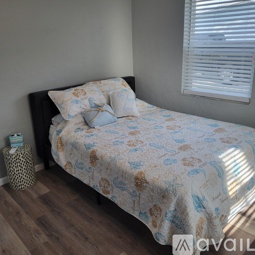 A bed with a floral patterned comforter and pillows is in a room with wooden flooring and a window with blinds.