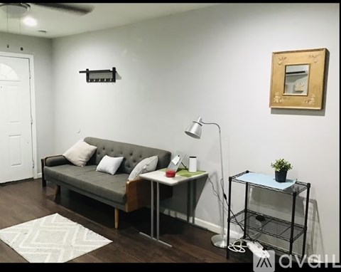 A living room with a grey couch, a small table with a lamp and a plant on it, and a picture frame on the wall.