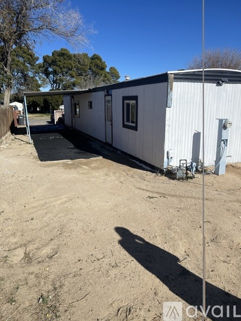 A white mobile home sits in a sandy lot with trees in the background.