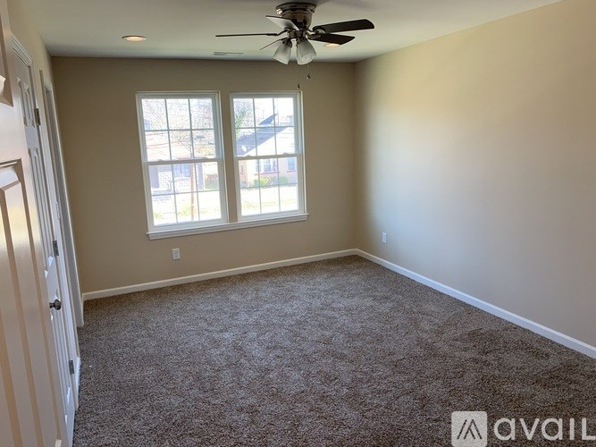 A room with a carpeted floor, a ceiling fan, and a window with a view of a house across the street.