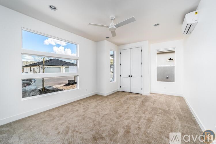 A room with a carpeted floor, a ceiling fan, and a window showing a view of a house and trees.