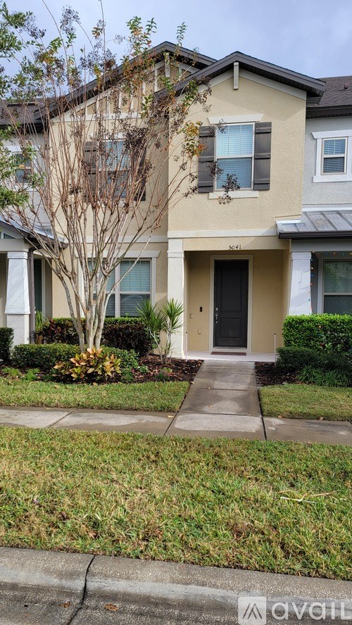 A beige house with a black door and a tree in front.