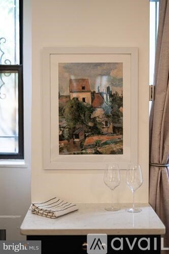 A painting of a village scene is displayed on a wall above a small table with two glasses and a napkin holder.