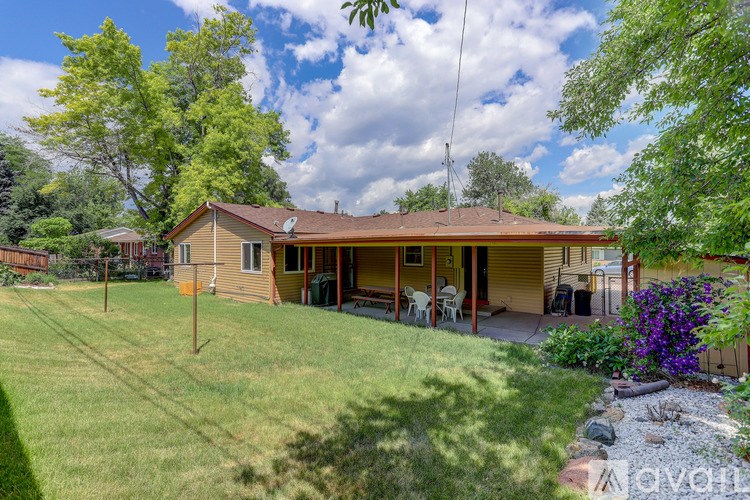 A house with a brown roof and a yellow wall is surrounded by a green lawn and trees.