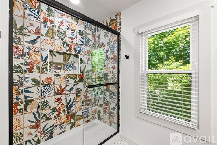 A bathroom with a floral tiled shower and a window with blinds.