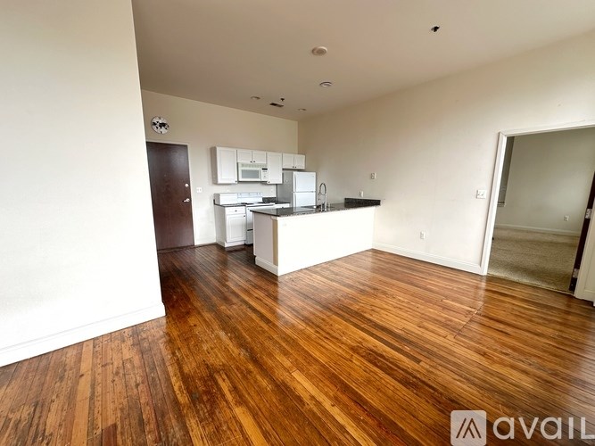 A room with wooden floors and a kitchen area in the background.