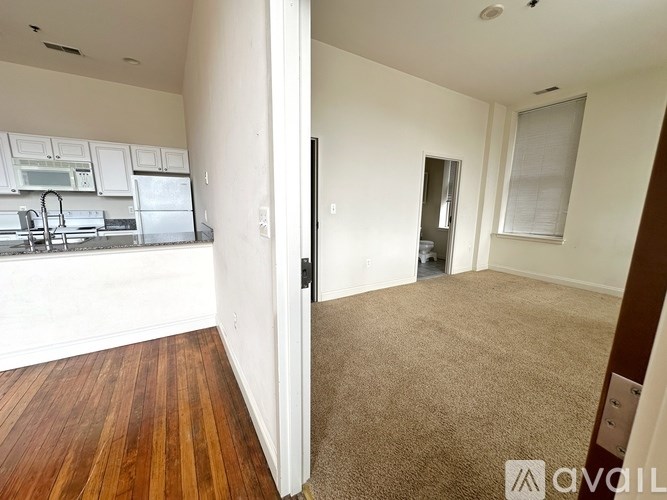 A room with a carpeted floor and a wooden floor leading to a kitchen area.