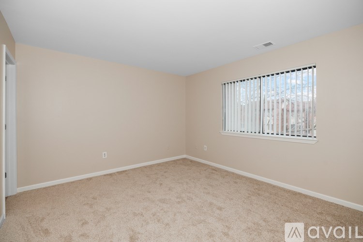 A room with beige walls and carpeting, a window with blinds, and a door on the left.