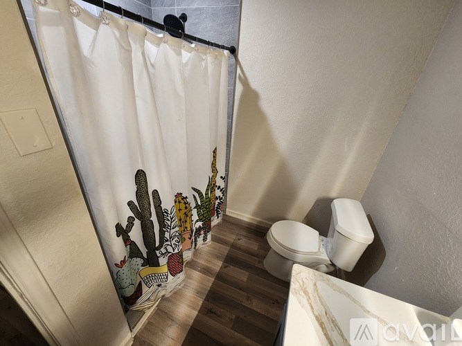 A bathroom with a toilet, a shower curtain with cacti on it, and a wooden floor.