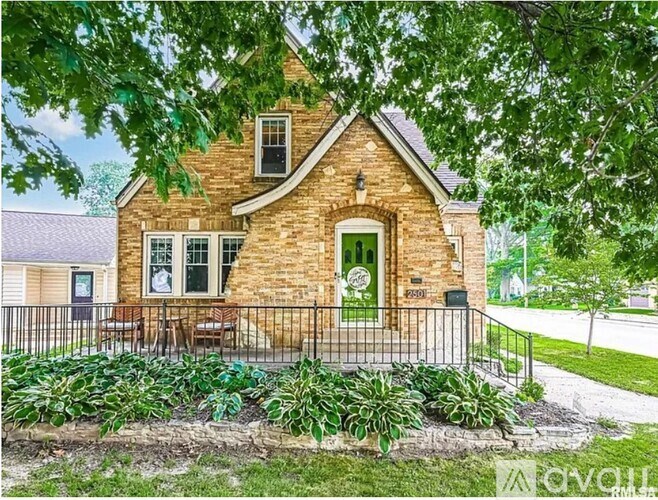 A small brick house with a green door and windows surrounded by a black fence and green plants.