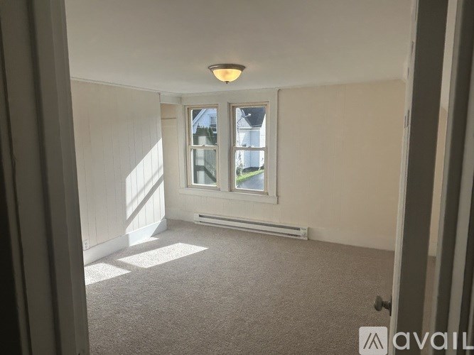 A room with a carpeted floor, a window with a view of a building outside, and a radiator on the wall.