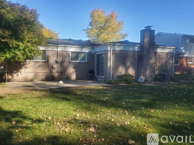A house with a brick exterior and a chimney is surrounded by a grassy lawn with fallen leaves.