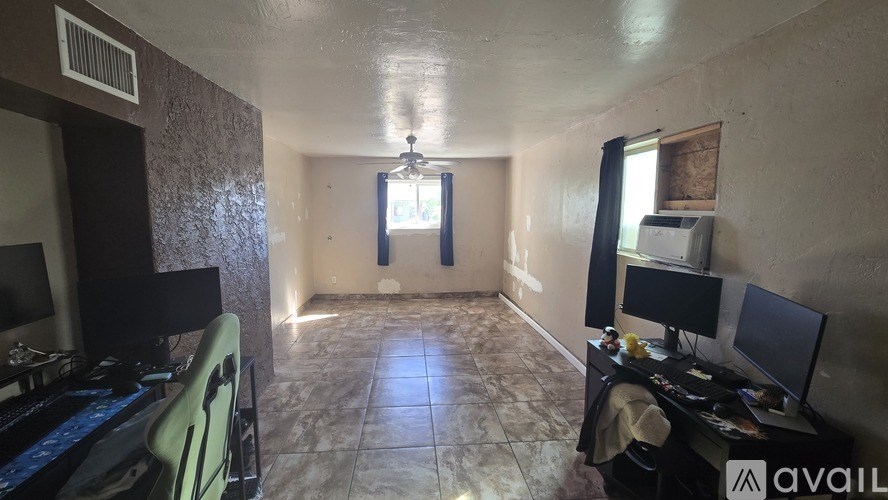 A room with a desk and computer setup in the foreground and a window with a view of the sky in the background.