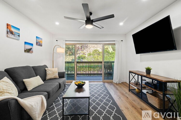 A living room with a grey sofa, a coffee table, a ceiling fan, and a flat screen TV mounted on the wall.