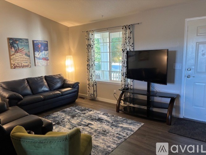 A living room with a black leather couch, a green armchair, a wooden rocking chair, a flat screen TV, and two posters on the wall.