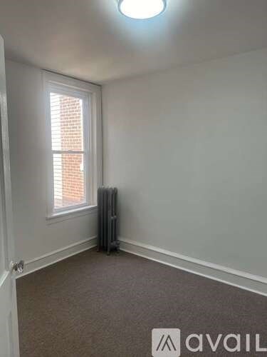 A room with a carpeted floor, a window with blinds, and a wall-mounted heater.