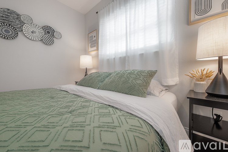 A bed with a green patterned blanket and a white pillow is in the foreground of a room with a lamp and wall art.
