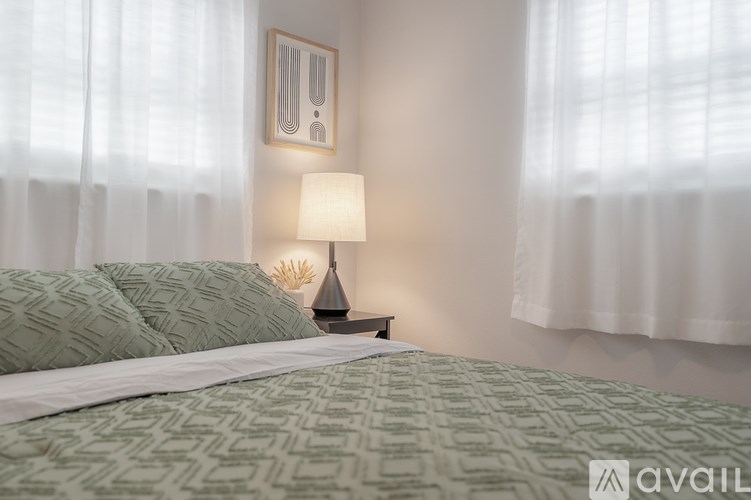 A bed with a green patterned comforter and a white pillow is in front of a window with white curtains.