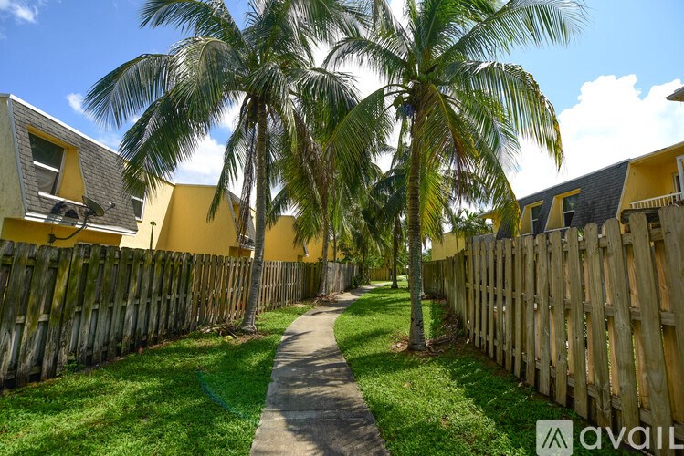A pathway leads through a grassy area between two wooden fences with palm trees in the background.