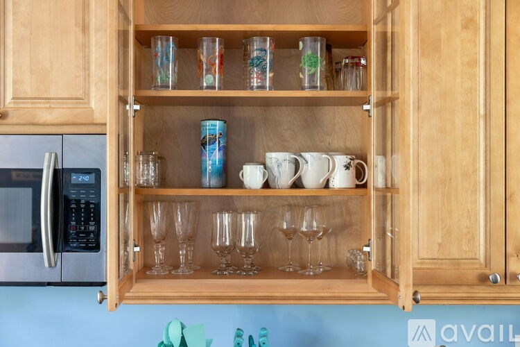 A kitchen cabinet with glass shelves holding cups, glasses, and a can.