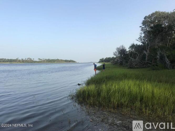 A person is standing on the shore of a calm body of water, holding a stick.
