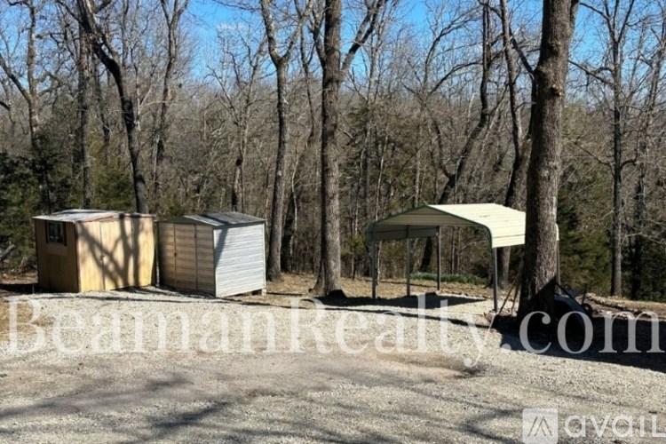 A real estate sign is displayed in front of a wooded area with two buildings.