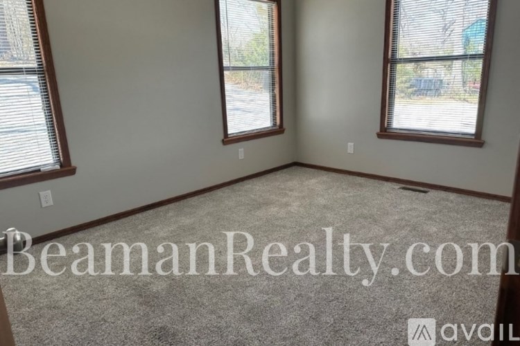 A room with two windows and a carpeted floor is available for rent.
