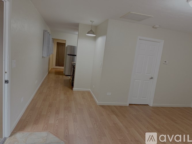 A room with wooden flooring and white walls, with a door on the right and a kitchen area in the background.