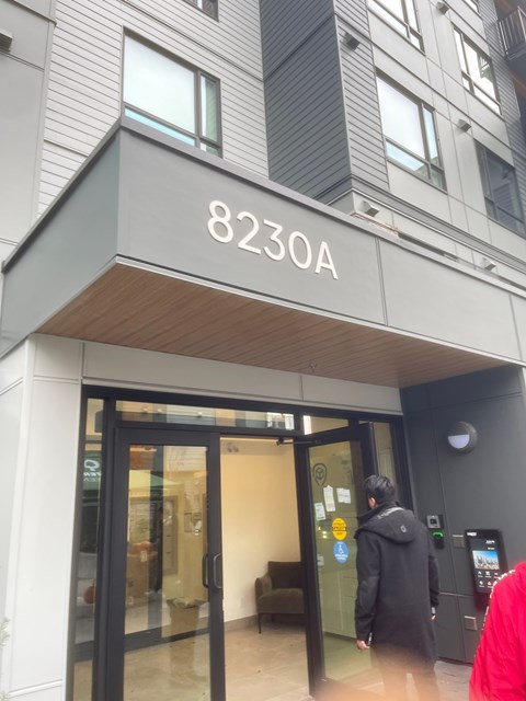 A person in a red jacket is walking past a building entrance with the number 8230A on it.