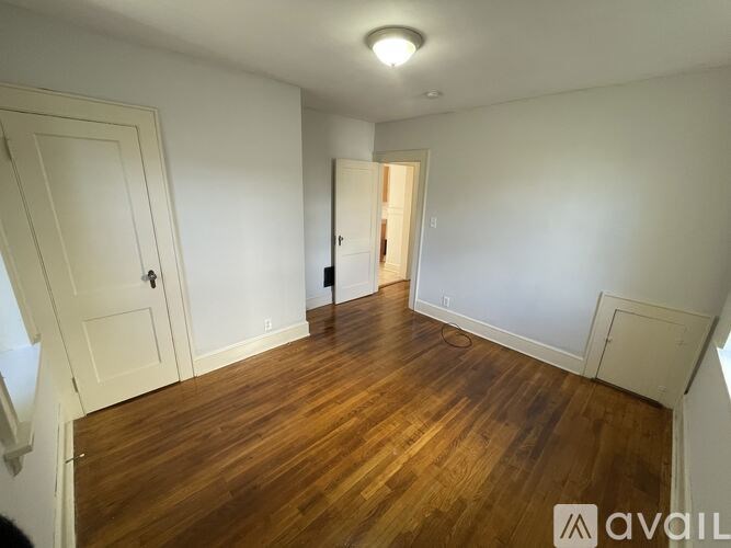 A room with wooden floors and white walls, with a door on the left and another door in the distance.